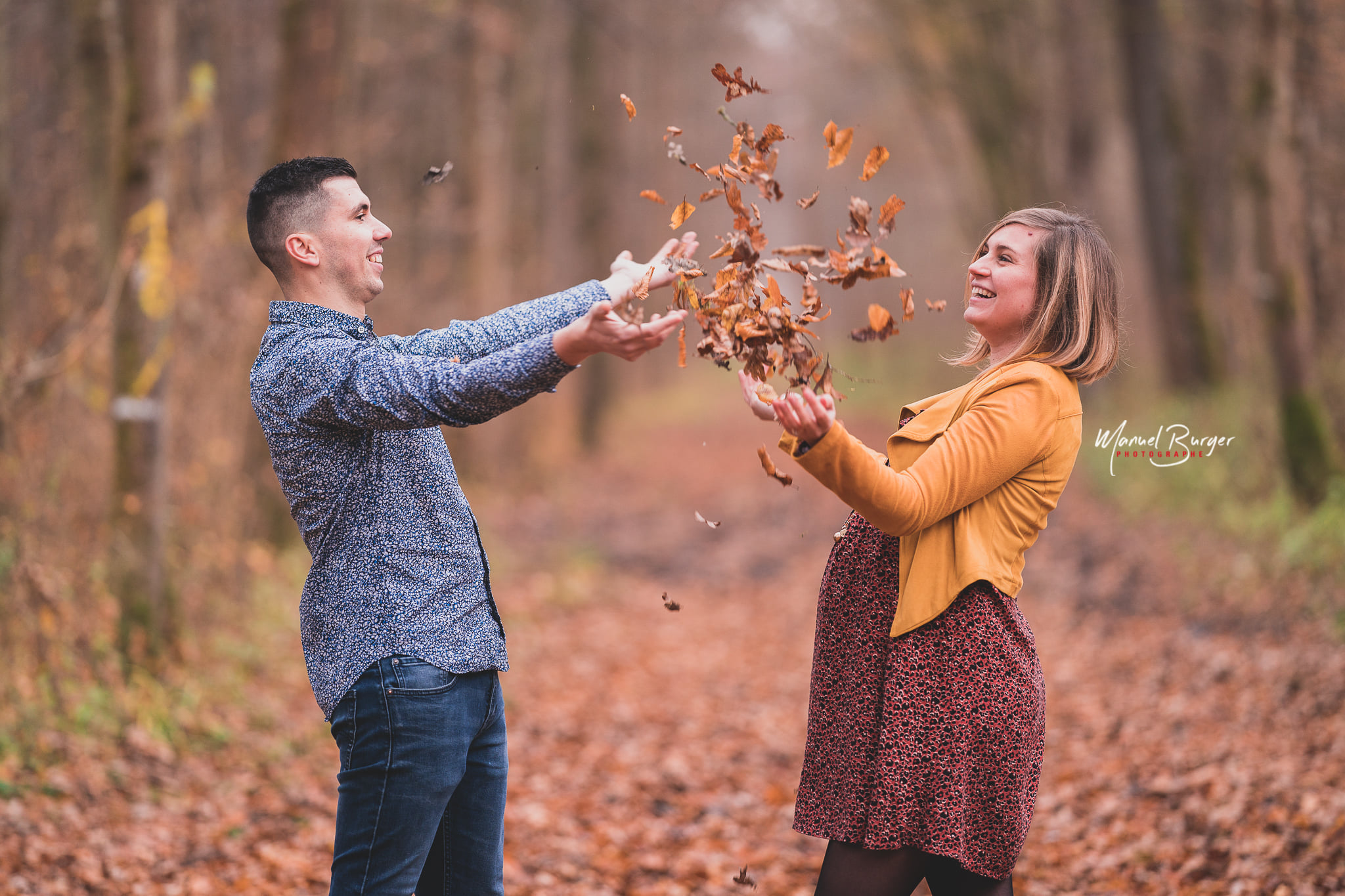 Éclat de rire d'un couple jetant des feuilles mortes en l'air pendant leur joyeuse séance photo de grossesse au cœur de l'automne à Sélestat.