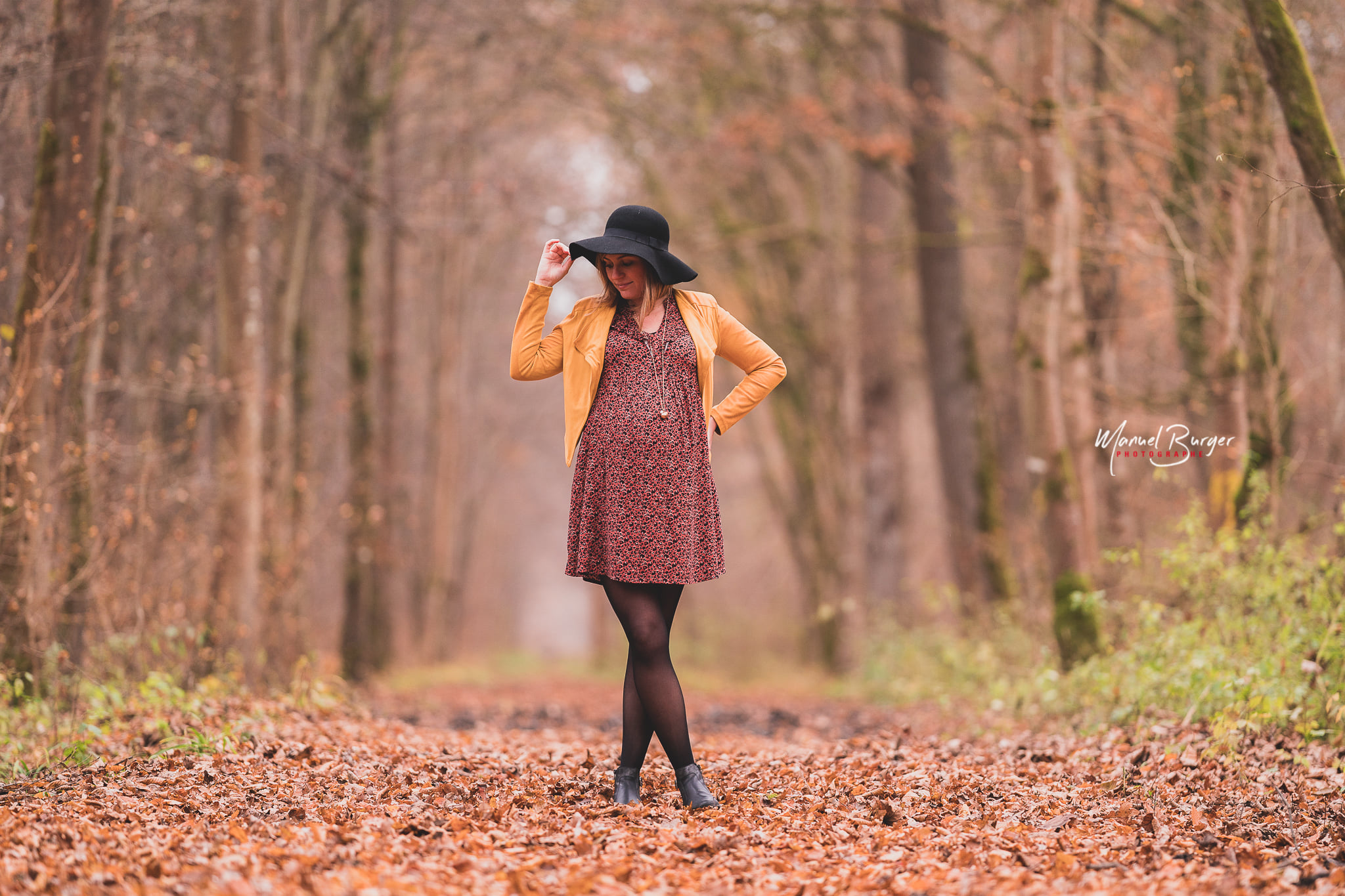 Femme enceinte élégante portant un chapeau noir, posant sur un chemin couvert de feuilles mortes dans la réserve de l'Illwald en Alsace.